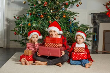 Noel kıyafetleri ve Noel Baba şapkaları giymiş üç çocuk, süslenmiş bir Noel ağacının önünde dizlerinde hediyelerle poz veriyorlar.