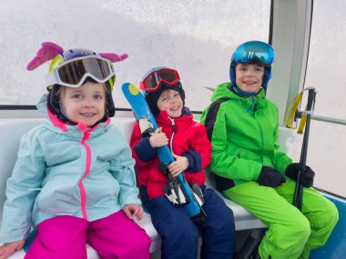 Bir grup mutlu çocuk renkli kayak kıyafetleri giyip teleferiğe binip, sıcak bir şekilde gülümsüyorlar.