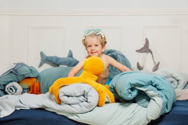 Çeşitli doldurulmuş deniz hayvanlarıyla dolu bir yatakta şnorkelli bir çocuk parlak turuncu pelüş bir oyuncak tutuyor.