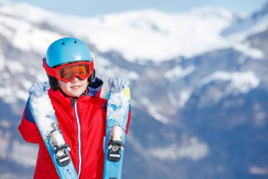 Kırmızı ceketli bir çocuk karda diz çöker, kar kaplı tepe manzaralı bir çift kayağı sıkar, kış aktivitesi portresi