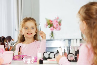 Kıvırcık saçlı bir kız rimel tutuyor, makyaj masasında makyaj yapıyor, kozmetik aletlerle çevrili, pembe çiçekler arka planı süslerken.