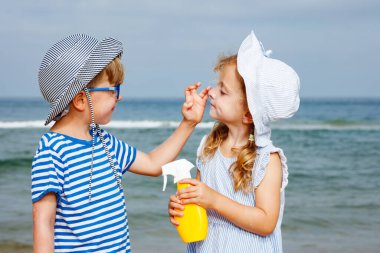 İki çocuk plajda, kız aile tatilinde elinde bir şişeyle kızın yüzüne güneş kremi sürüyor.