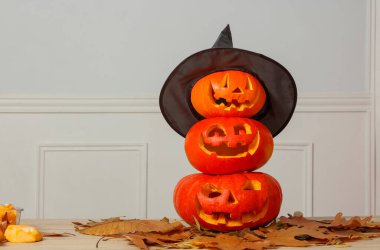 Three carved pumpkins are stacked on top of each other, wearing a black witch hat, surrounded by autumn leaves on a wooden table clipart