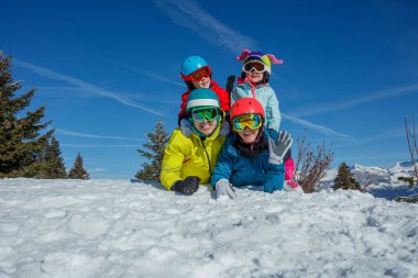 Çocukların ve ailelerinin parlak kayak kıyafetleri içinde karla kaplı tepelerin manzaralı manzarasıyla karın üzerinde yattıkları eğlenceli bir kış sahnesi.