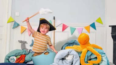 Siyah korsan şapkalı çocuk, elinde ev yapımı bayrakla içi doldurulmuş deniz oyuncakları ve canlı afişlerle dolu bir odada oynuyor.