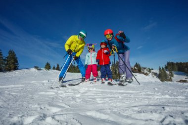 Aydınlık bir kış günü, bir aile canlı bir kayak elbisesi içinde karlı bir tepede kayak yapmaya hazırlanıyor.