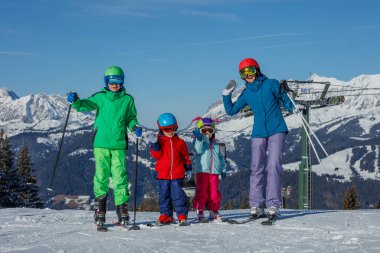 Güneşli bir günde, kış tatillerinde dağlarda kayak yaparken kameraya gülümseyen ve el sallayan dört kişilik bir aile.