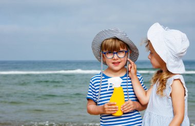 Şapka takan ve çizgili kıyafet giyen iki çocuk, aile tatillerinde ellerinde sarı bir sprey şişeyle denizin kenarında duruyorlar.