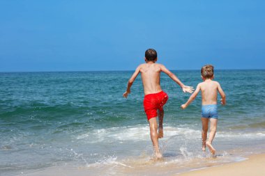 Denize doğru koşan iki çocuk görülüyor, kıyı şeridinde su sıçratıyor, yaz tatilleri eğlencelidir.