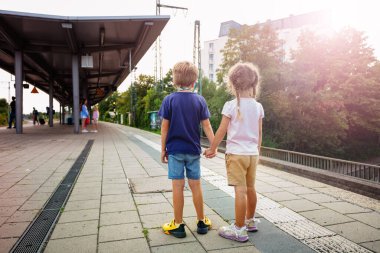 Çocuklar, tren istasyonunda beklerken, yaz tatili boyunca el ele tutuşurken, hareket eden trenleri gözlemlerler.