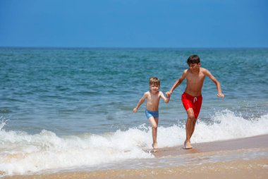 İki erkek kardeş, kumsalda mutlu bir şekilde koşuyorlar ve arkalarında dalgalar var.