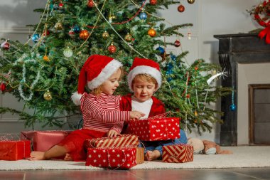 Süslü bir Noel ağacının altında Noel Baba şapkalı iki çocuk hediyelerini açıyor. Mutlu ve nişanlı görünüyorlar.