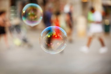 Renkli bir sahneyi yansıtan sabunlu bir baloncuğun yakın plan görüntüsü. Arka planda bulanık figürler yürüyor.