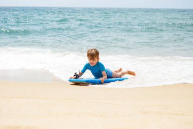 Kumsalda, küçük bir çocuk sörf tahtasıyla oynarken dalgalar yavaşça kumlara vuruyor.