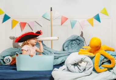 Korsan şapkası takmış bir kız, büyük bir kovanın içinde oturmuş kağıt rulo bir teleskoptan bakıyor, arkasında renkli flamalarla denize açıldığını hayal edin.