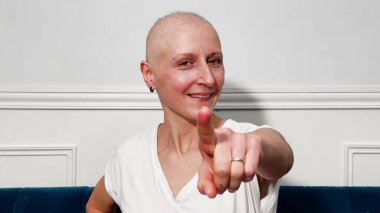 Mavi bir kanepede duvara yaslanmış beyaz gömlekli, gülümseyen kel kadın kamerayı işaret ediyor.