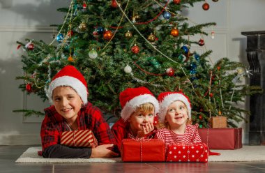 Noel Baba şapkalı üç neşeli çocuk, neşeli bir şekilde Noel ağacının yanında oturmuş hediyelerini tutuyorlar.
