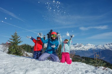 Anneleri olan çocuklar, parlak ve renkli kışlık giysiler giyiyorlar, güneşli bir günde dağlarda tatil yaparken neşeyle havaya kar atıyorlar.