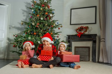 Bir grup kırmızı Noel Baba şapkalı çocuk Noel ağacının yanında oturmuş, ellerinde hediyelerle heyecanlı bir şekilde gülümsüyorlar.