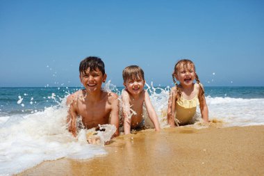 Sahilde sürünen gülen çocuklar, arka planda deniz, köpüklü dalgalar etrafa sıçrayan çocuklar, heyecan çığlıkları.