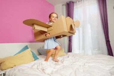 Yatağında duran genç bir kız renkli renkli bir yatak odasında karton bir uçakla oynarken ışık saçıyor.