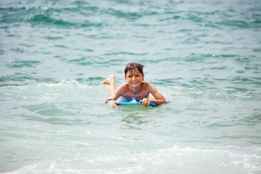 Neşeli bir çocuk mavi bir sörf tahtasının üzerinde yatıyor, dalgaların arasında süzülüyor ve yaz tatillerinde gülümsüyor.