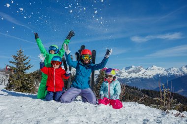 Anneleri ile birlikte renkli kış giysileri içinde bir grup çocuk güneşli bir günde açık mavi gökyüzüne kar yağdırıyor.