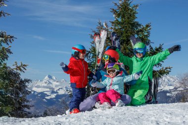 Güneşli bir kış gününde, bu aile renkli kayak kıyafetleriyle bir fotoğraf için ara verir. Arkalarında kayaklar ve resimli dağlar vardır.