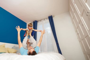 Bir baba ile oğlunun düzenli bir yatak odasında kep takıp çocuğu tavana kaldırması arasında neşeli bir an.