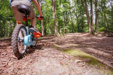 Küçük çocuk ormanda bir patika boyunca bisiklet sürüyor, günlük şort giymiş, bacakları ve tekerlekleri alçak açıdan görüyor.