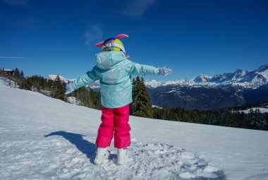 Neşeli miğferli kayakçı kız kollarını dağlara uzatmış, arkadan manzaralı.