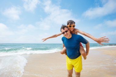 Bir baba ve genç bir oğul kumsalda eğleniyor, babasının sırtında binen çocukla, okyanus dalgaları içeri girerken