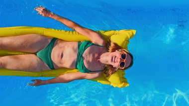 A woman relaxes on a yellow inflatable raft in the clear blue waters of a swimming pool, wearing a green bikini and orange sunglasses, enjoy vacations clipart