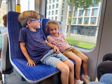 İki çocuk tramvayın içinde mavi bir koltukta oturuyorlar, arka planda pencereden görünen bir şehir manzarası var.