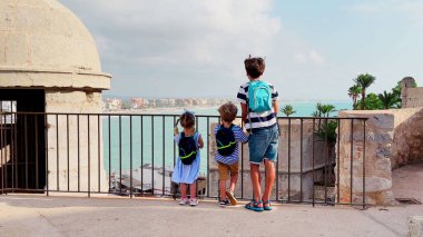 Üç çocuktan oluşan bir grup, Peniscola Kalesi 'nin duvarlarından okyanus manzaralı bir şehir alanını görebilmek için metal bir bariyerin ötesine bakıyorlar.