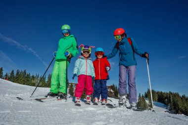 Canlı kayak kıyafeti giymiş bir grup insan açık mavi gökyüzünün altında kayak yapmaya hazır, karlı bir tepede duruyor.