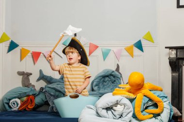 Siyah korsan şapkalı çocuk, elinde ev yapımı bayrakla içi doldurulmuş deniz oyuncakları ve canlı afişlerle dolu bir odada oynuyor.