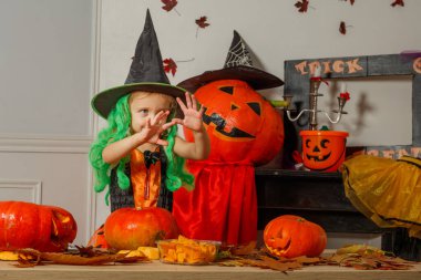 Yeşil bukleli cadı kıyafeti giyen küçük bir kız, siyah şapka oyulmuş balkabakları ve şenlikli dekorun önünde neşeli hareketler yapıyor.
