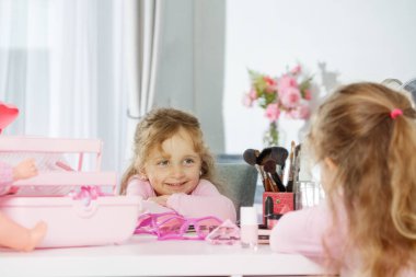 Kozmetik eşyalarla dolu bir makyaj masasında oturan pembe elbiseli mutlu bir kız aynaya bakıyor.