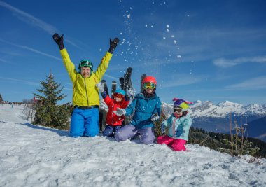 Canlı bir grup çocuk ve ebeveyn, renkli kışlık ceketler içinde, neşeyle yukarıya kar fırlatıyorlar, muhteşem karlı dağların yanına, güneşli bir gökyüzü manzarayı hazırlıyor.