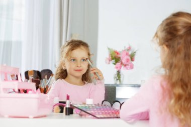 Küçük bir kız makyaj yapıyor makyaj masasında otururken aynanın önünde kozmetik malzemelerle dolu, arka planda pembe bir çiçek buketi.