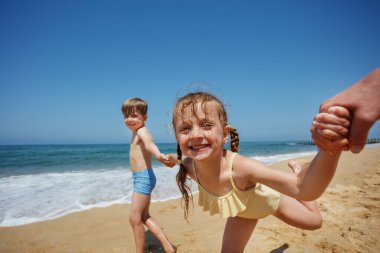 İki çocuk el ele tutuşuyor ve arka planda okyanus olan güneşli bir sahilde gülümsüyor.