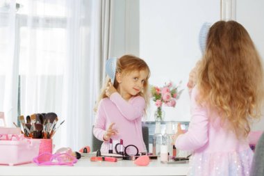 Açık pembe tişörtlü küçük bir çocuk saçlarını zevkle tarar, makyaj malzemeleriyle gösterişli bir yere oturur.