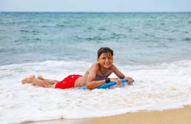 Kırmızı mayo giyen genç bir çocuk sörf tahtasında dalgaları bekliyor ve gülümsüyor.
