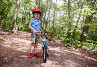 Kırmızı kasklı ve çizgili tişörtlü gülümseyen bir çocuk güneşli yaz ormanlarında bisikletinin yanında durup iz sürmeye hazırdır.