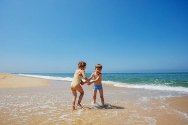 Genç bir çift, kız ve erkek kumlu sahilde birlikte koşarken, arkalarında deniz uzanırken görülüyorlar.