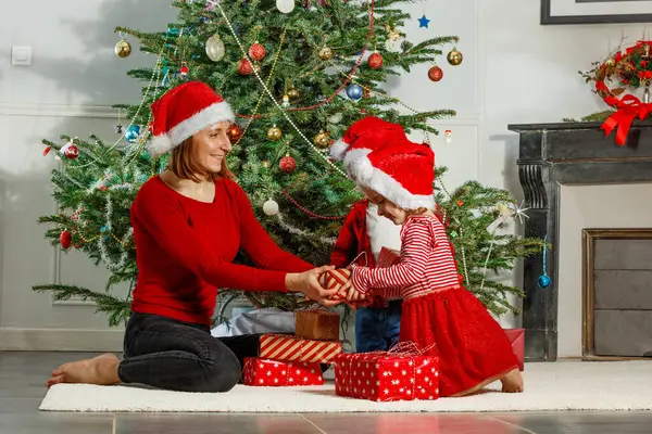 Kırmızı Noel Baba şapkalı bir anne ve çocuklarının güzel dekore edilmiş bir Noel ağacının yanında hediyeler açtığı neşeli bir tatil anı.