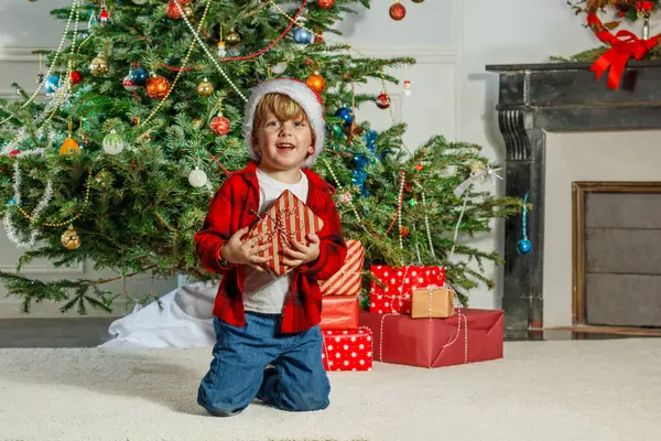 Noel Baba şapkası takan bir çocuk, renkli bir Noel ağacının önünde elinde hediyelerle neşeyle ışıldıyor.