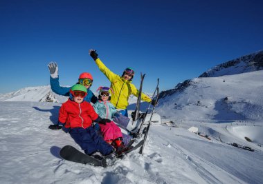 Dört kişi, renkli kayak kıyafetleri içinde, dağların panoramik manzarası ve yukarıdaki parlak mavi gökyüzü ile karla kaplı bir yamaçta rahatlar.