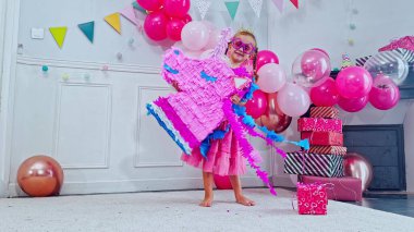 Elinde tek boynuzlu at pinatasıyla, kız oturma odasında duruyor pembe balonlar ve bayram flamalarıyla süslenmiş, bir kutlama hissi veriyor.